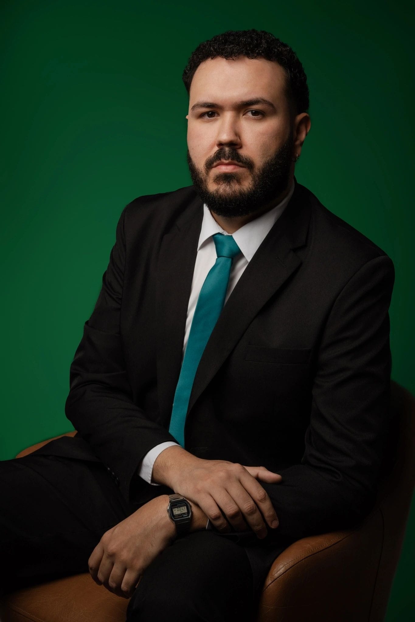 Clayton Medeiros, de terno preto e gravata azul-petróleo, sentado numa poltrona caramelo diante de um fundo verde.