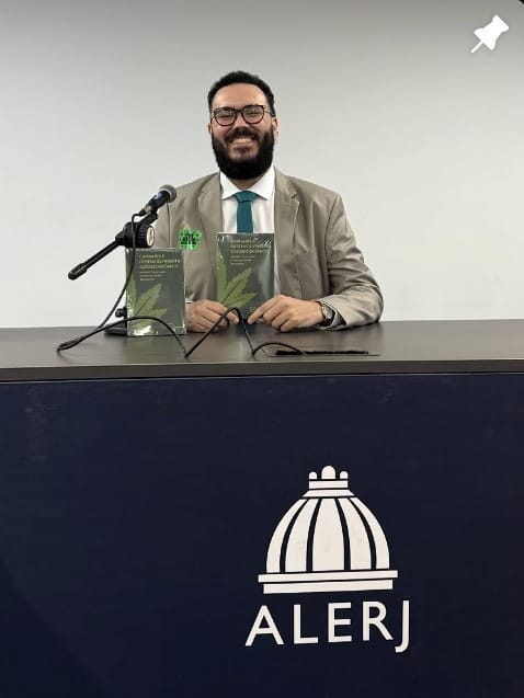 Clayton Medeiros sorrindo a uma mesa da Alerj, com dois livros à frente.